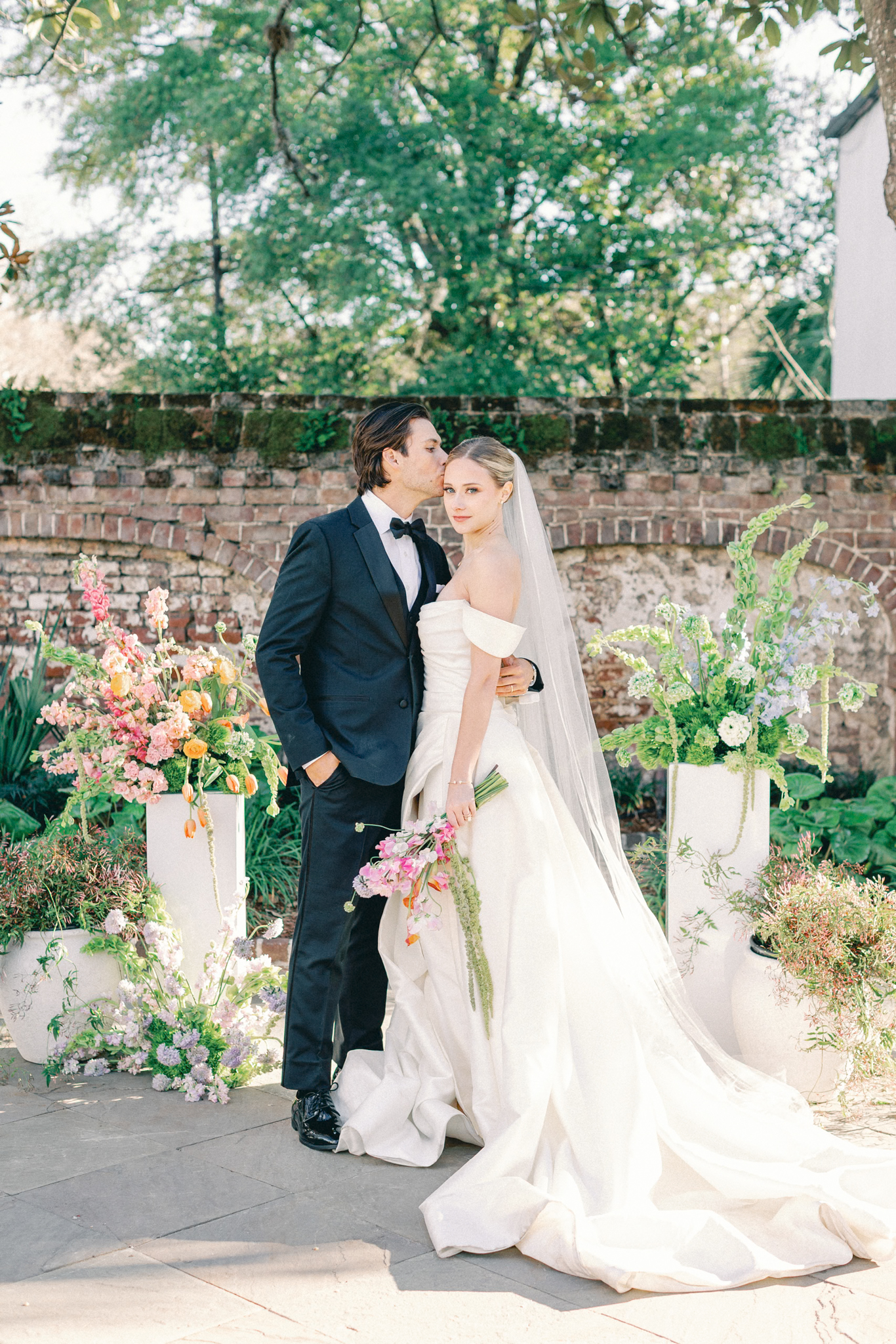Groom kisses his bride's forehead by a curved brick wall lined with tall spring florals — a walled-garden Charleston wedding venue