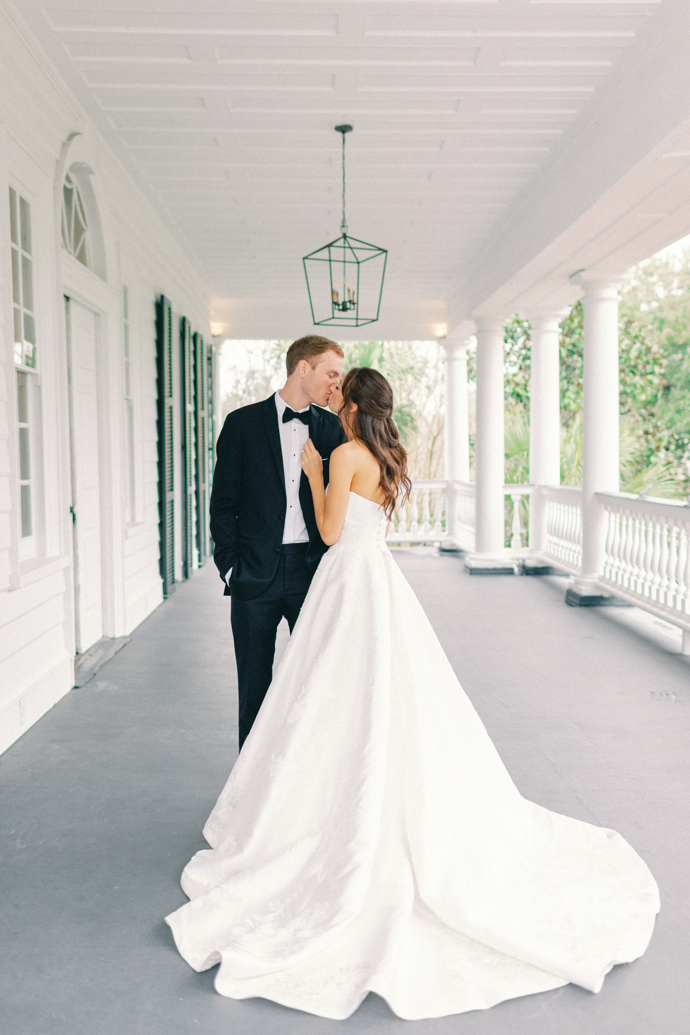 Couple kisses on a columned piazza beneath a green iron lantern — the porch of an elegant Charleston wedding venue, lowndes grove