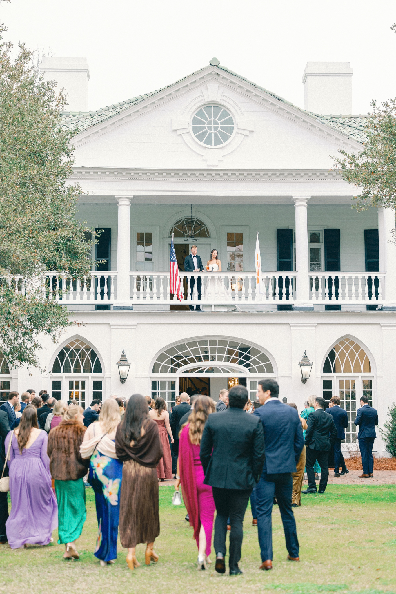 Guests gather on the lawn as a couple stands on the upper piazza of a white mansion — a riverside Charleston wedding venue