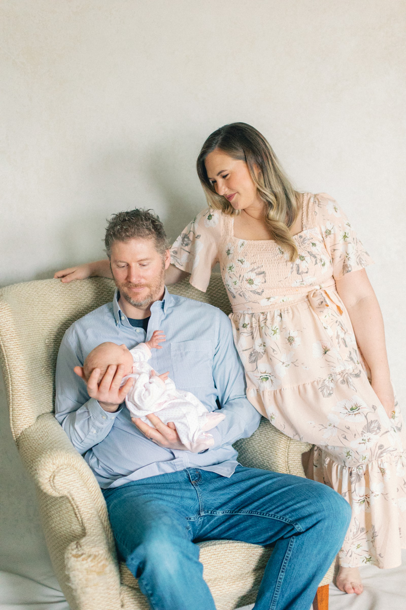 parents with newborn baby in front of fine art backdrop sitting on a chair