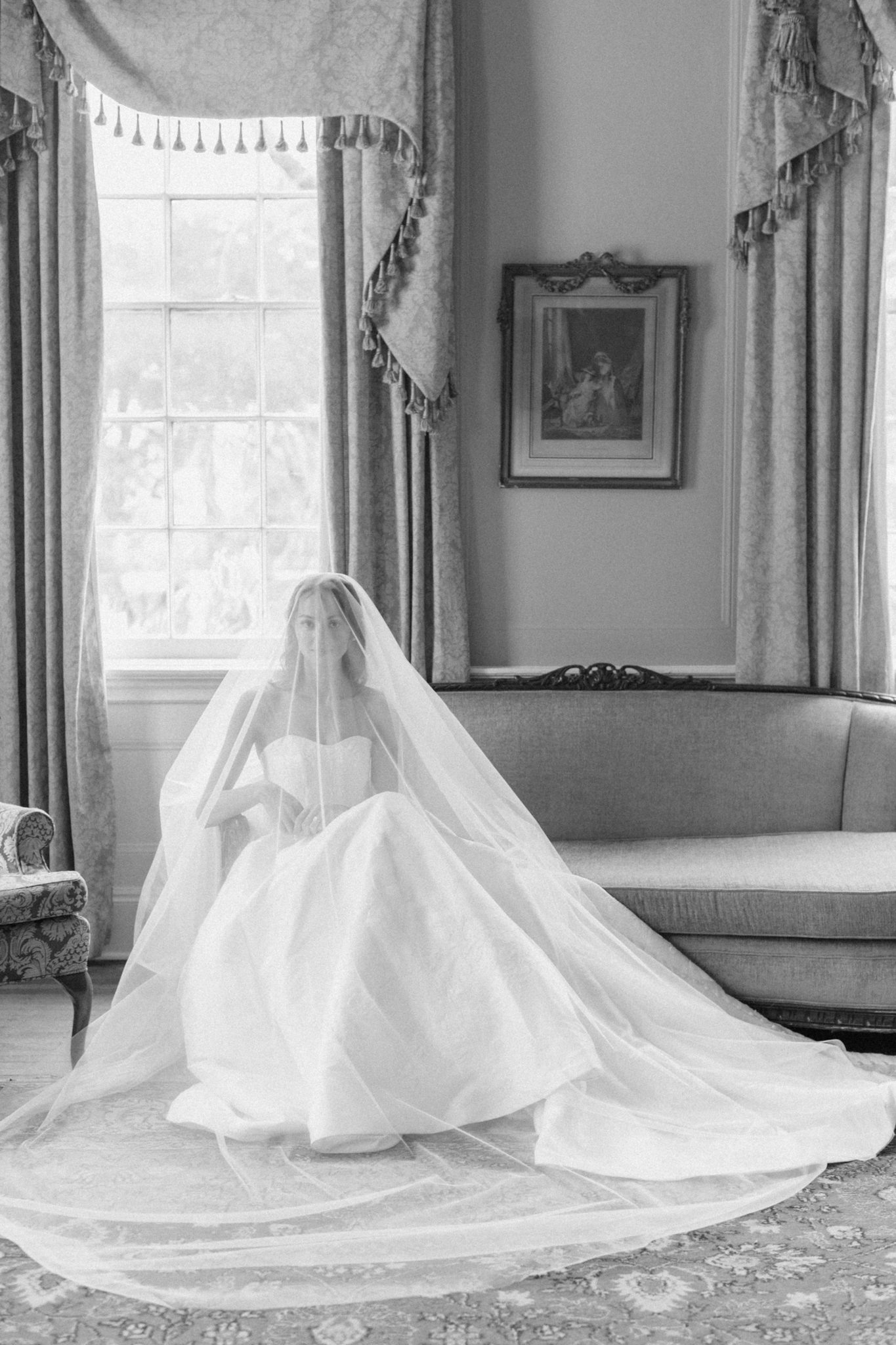 Black-and-white bridal portrait of a veiled bride seated by an antique sofa in a formal parlor with damask drapes and framed art, lowndes grove