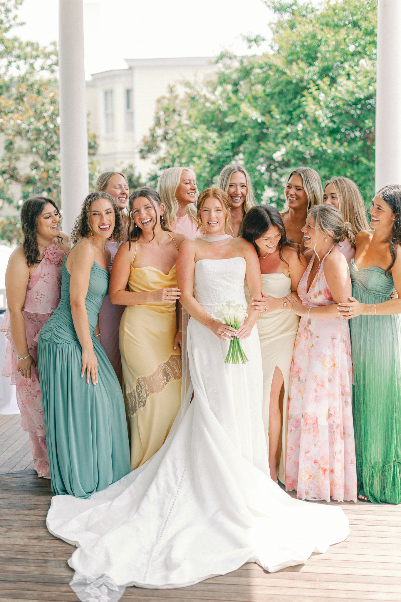 A bride laughs with her bridesmaids in mismatched pastel dresses on a piazza at a spring wedding in Charleston