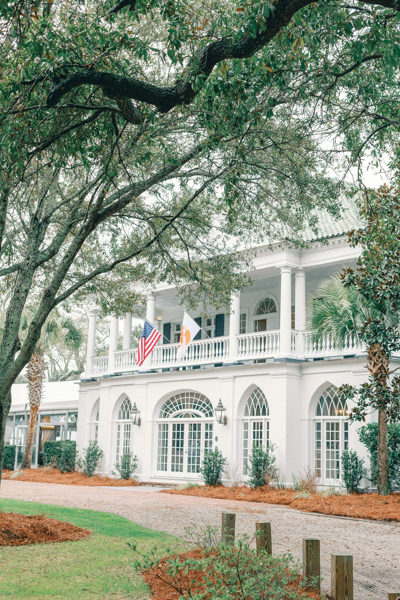 The white portico and arched windows of a waterfront estate flying an American flag under a live oak, a Charleston wedding venue