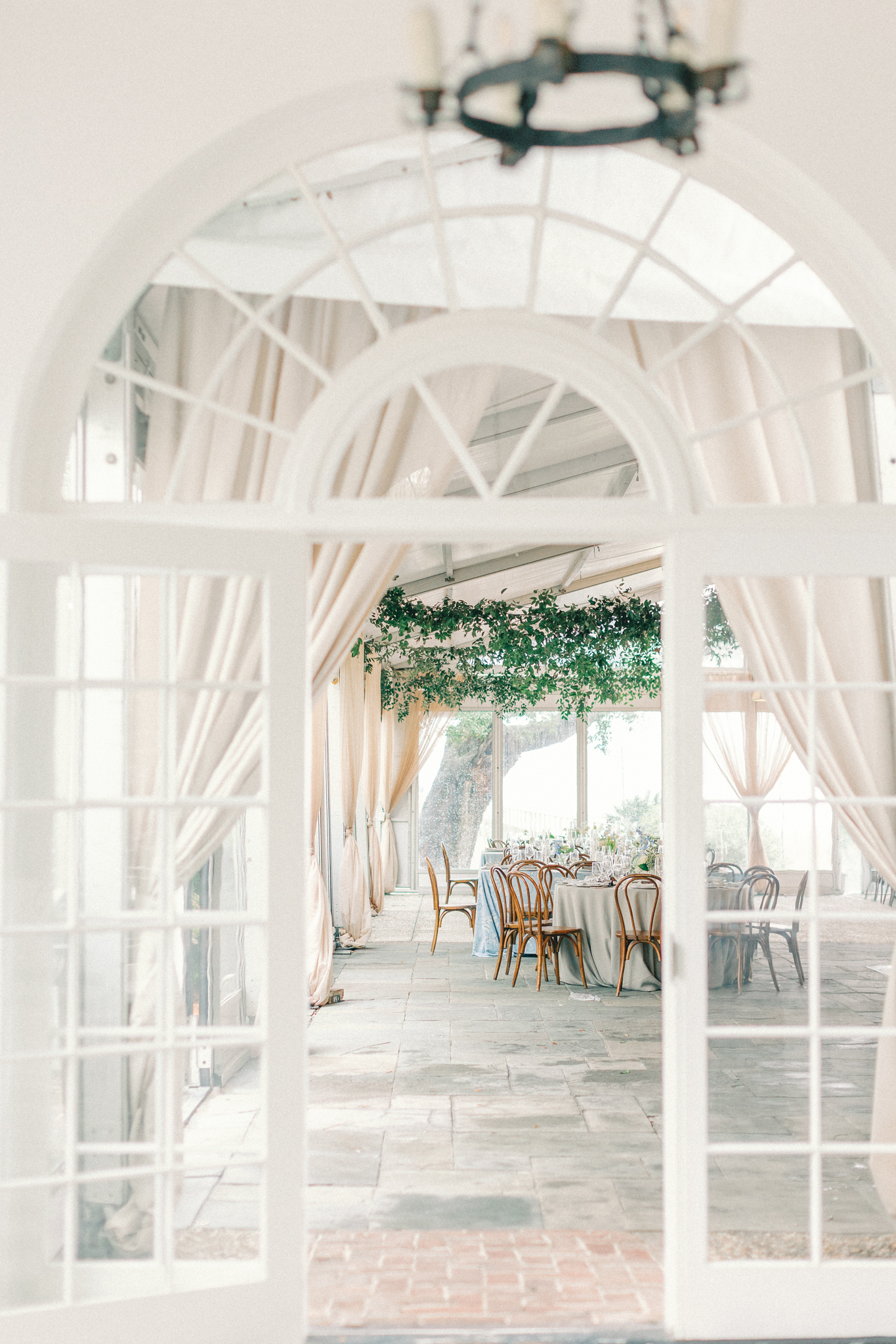 View through arched French doors into a reception with bentwood chairs, blue linens, and hanging greenery at a Charleston wedding venue