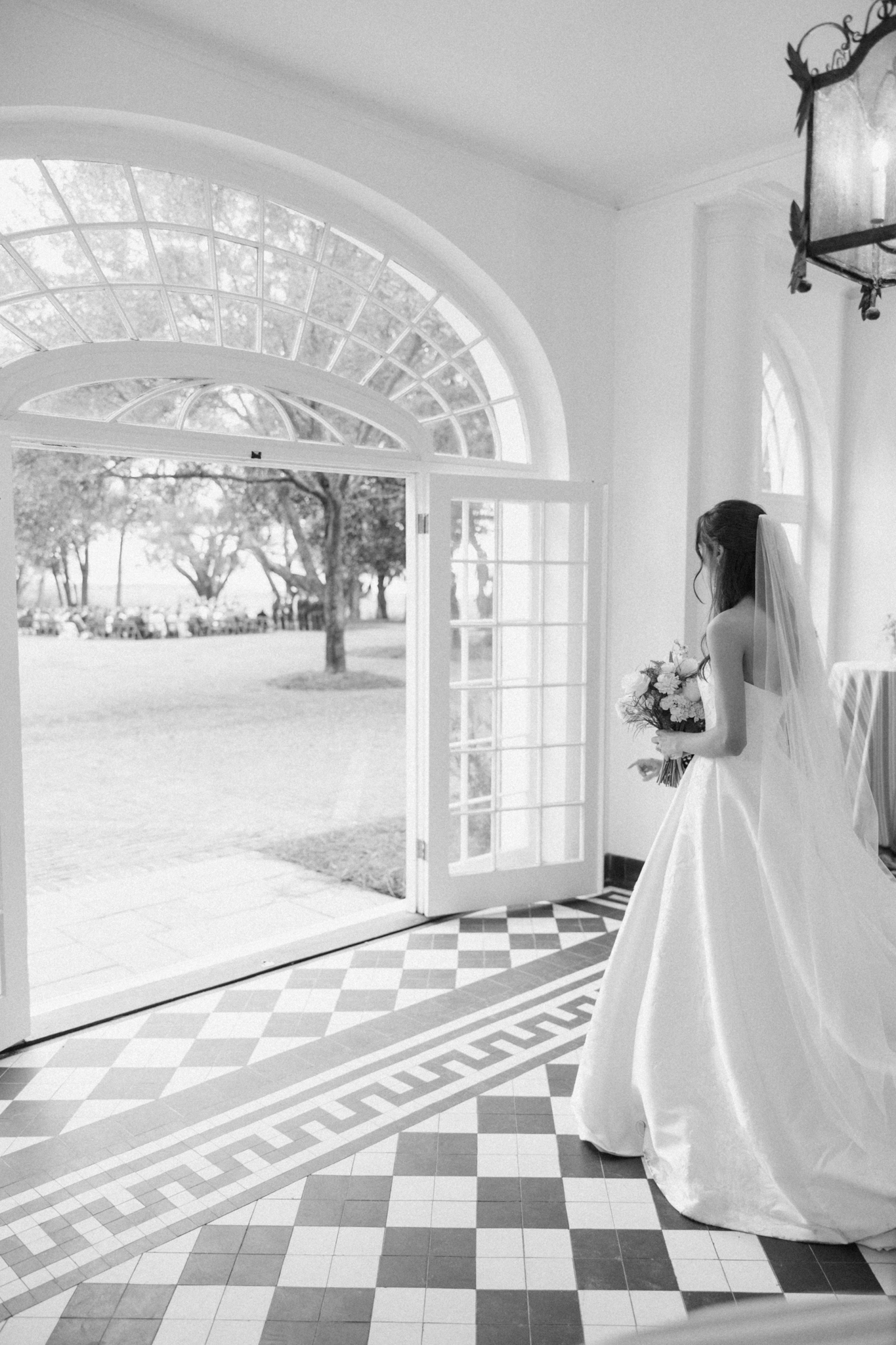 Black-and-white photo of a veiled bride in a loggia with a checkered marble floor looking out an arched door to the ceremony lawn