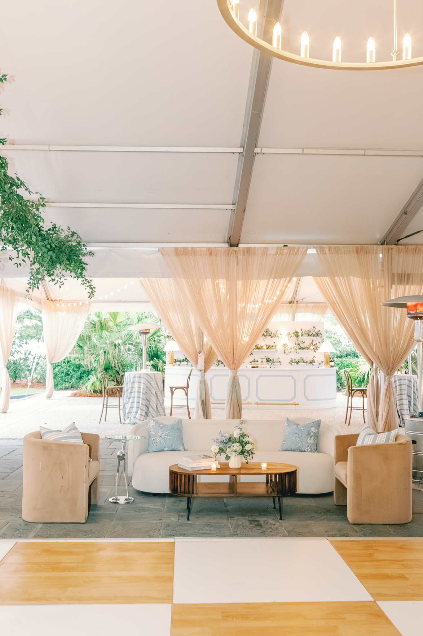 A tented lounge with blush drapes, a curved sofa, velvet chairs, and a bar beside a checkerboard dance floor at a Charleston wedding venue