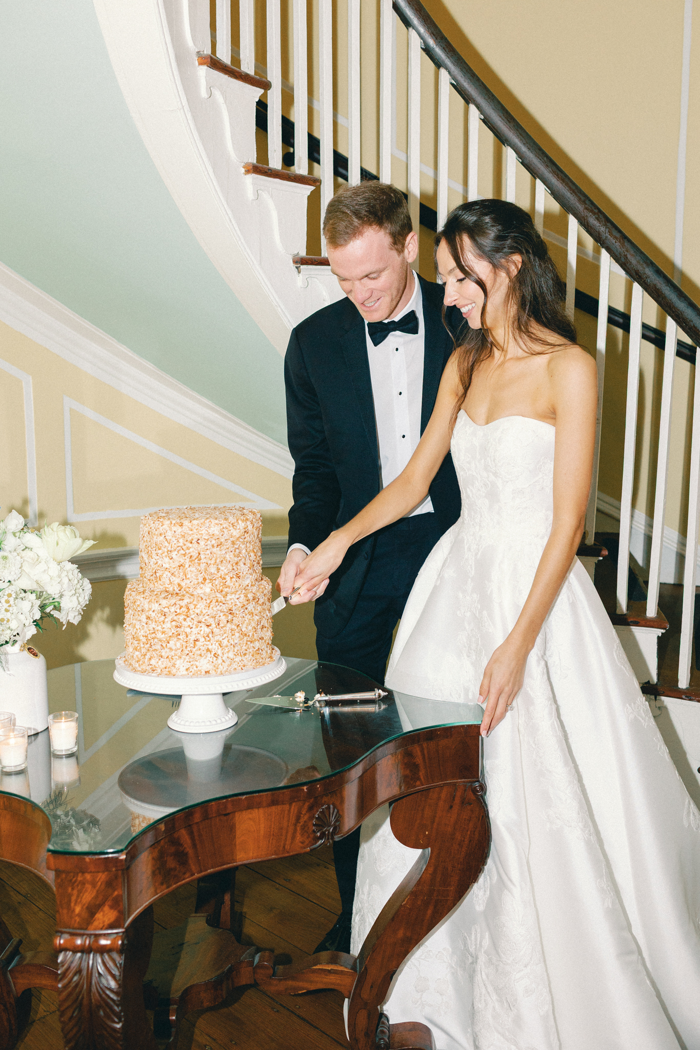 Bride and groom cut a textured two-tier cake on an antique table beside a curved staircase