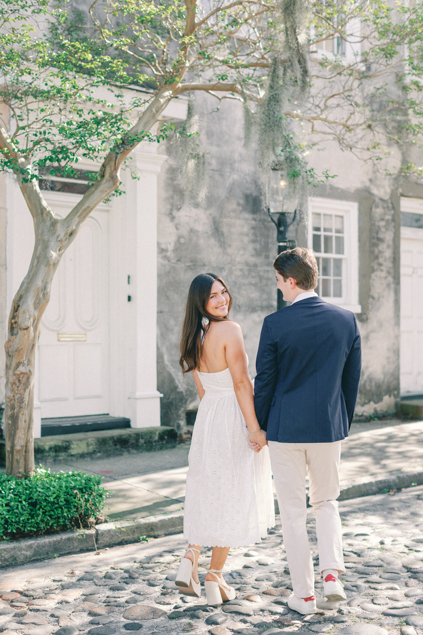Couple walking away on a cobblestone street, woman glancing back in a white eyelet dress, during Charleston engagement pictures