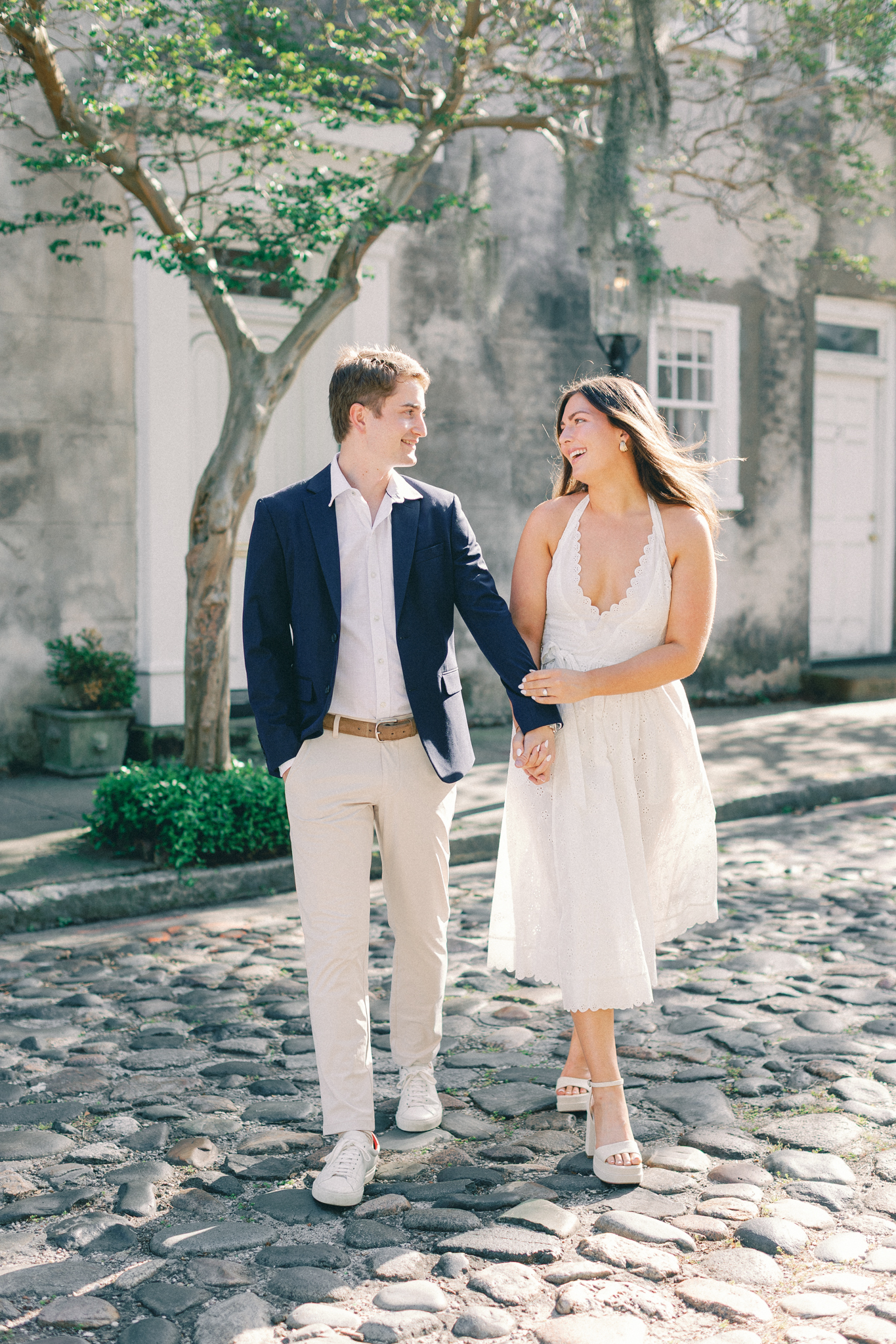 Couple in a navy blazer and white eyelet dress walking a cobblestone street for their Charleston engagement pictures
