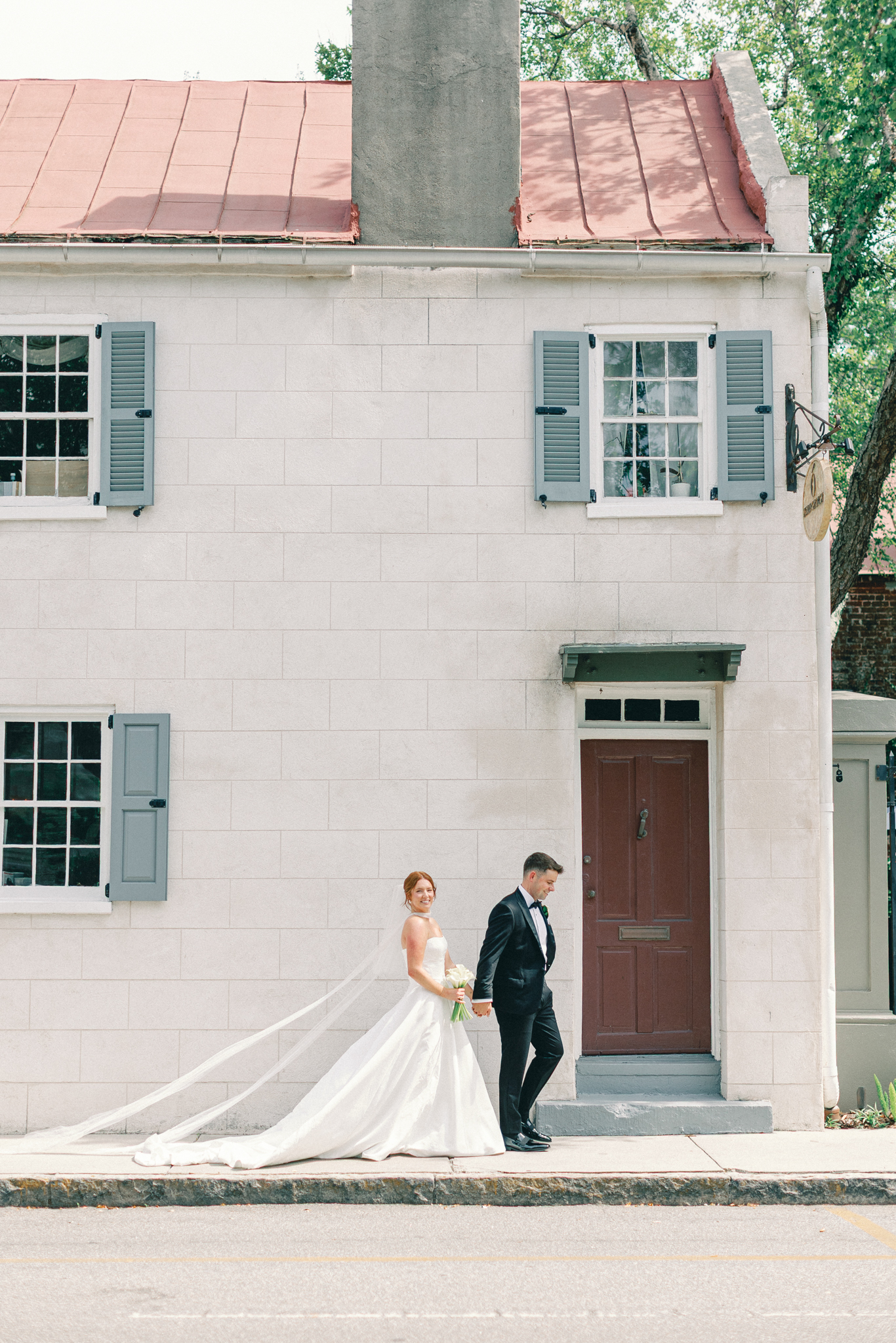 Bride and groom walking a downtown Charleston street past a historic home with a red tin roof and blue shutters