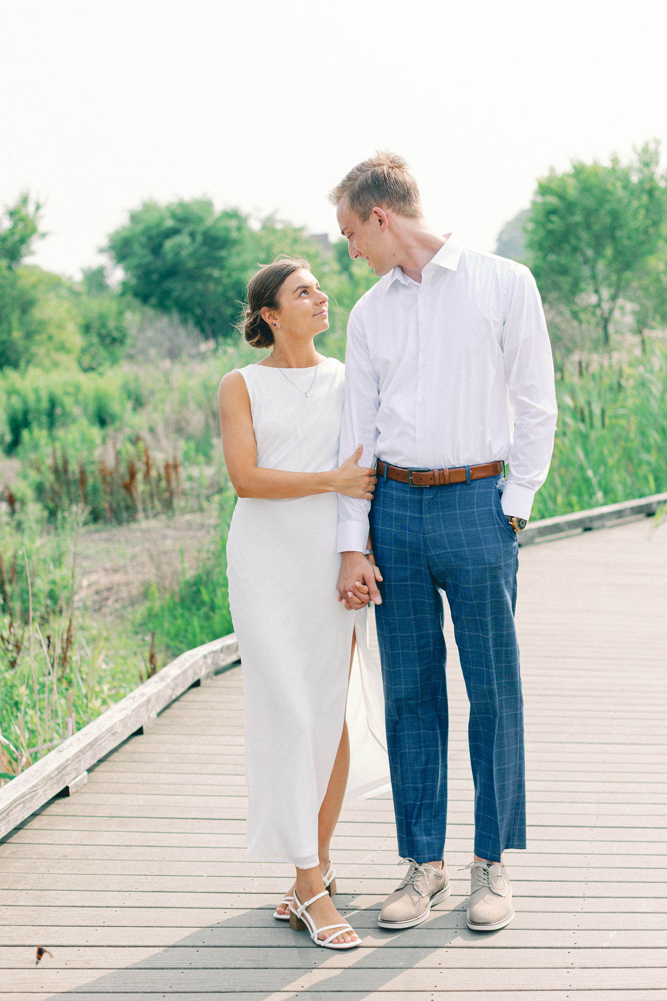 Emma and Caleb hold hands on a boardwalk through marsh grass during their Michigan engagement photos in Port Huron.