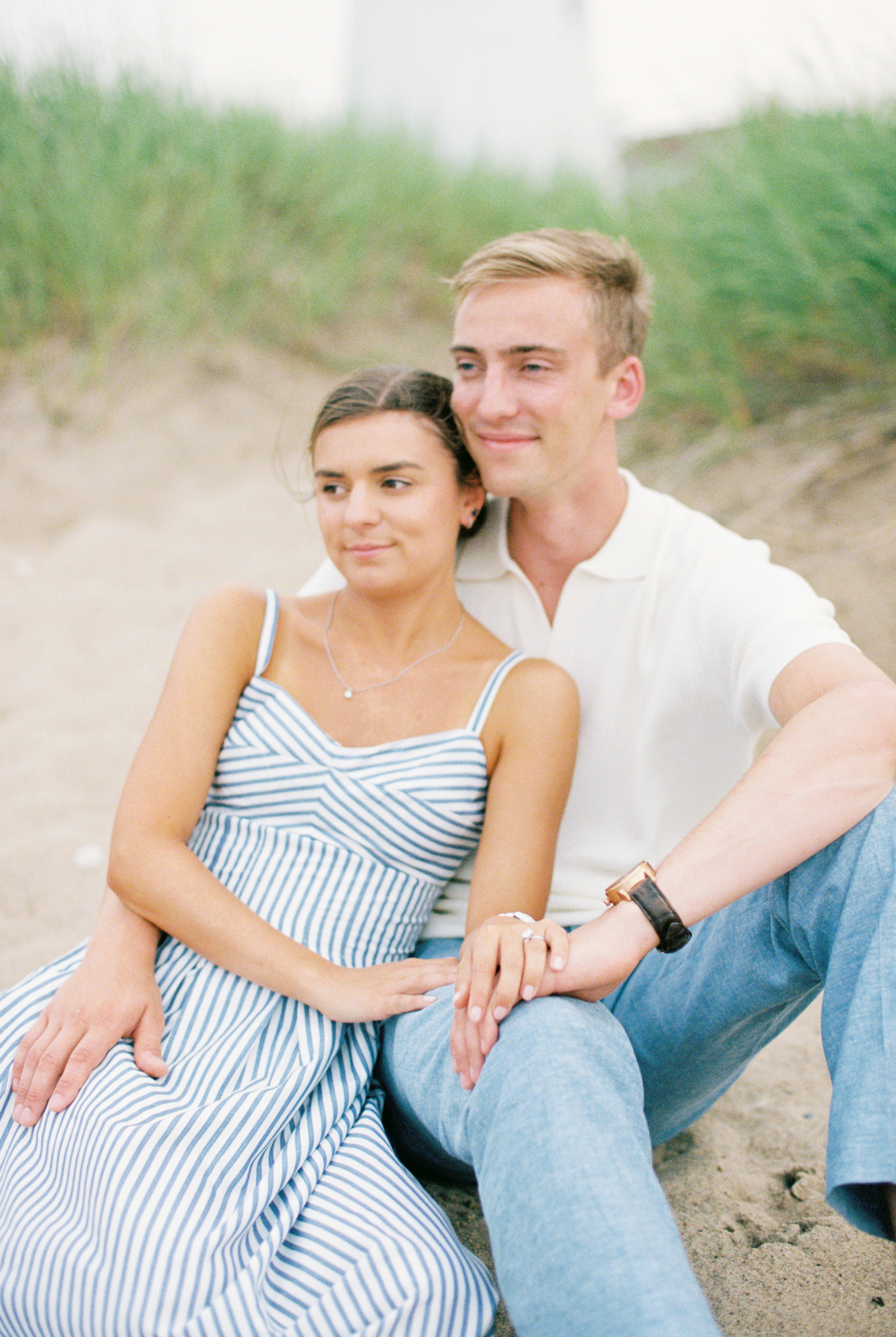 Emma leans against Caleb as they sit together in the sand during their Port Huron, Michigan engagement session.
