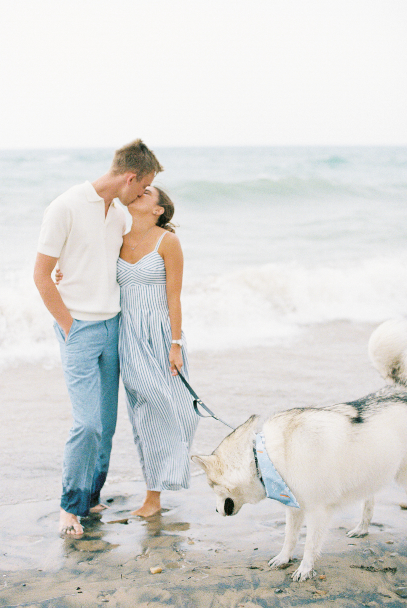 Emma and Caleb kiss on the beach as their husky sniffs the sand, Port Huron, Michigan engagement photography.