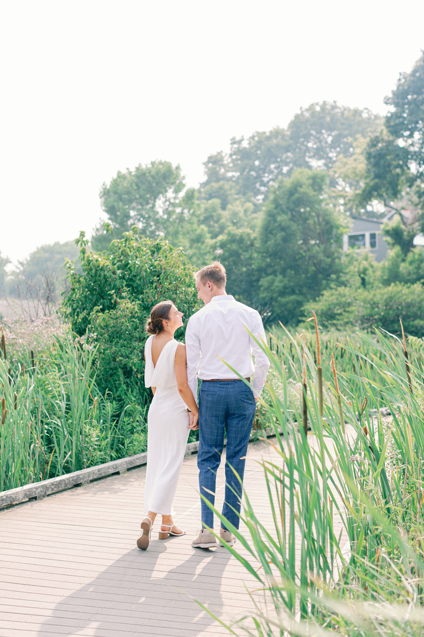 Emma and Caleb glance at each other over their shoulders while holding hands on a Port Huron, Michigan boardwalk.