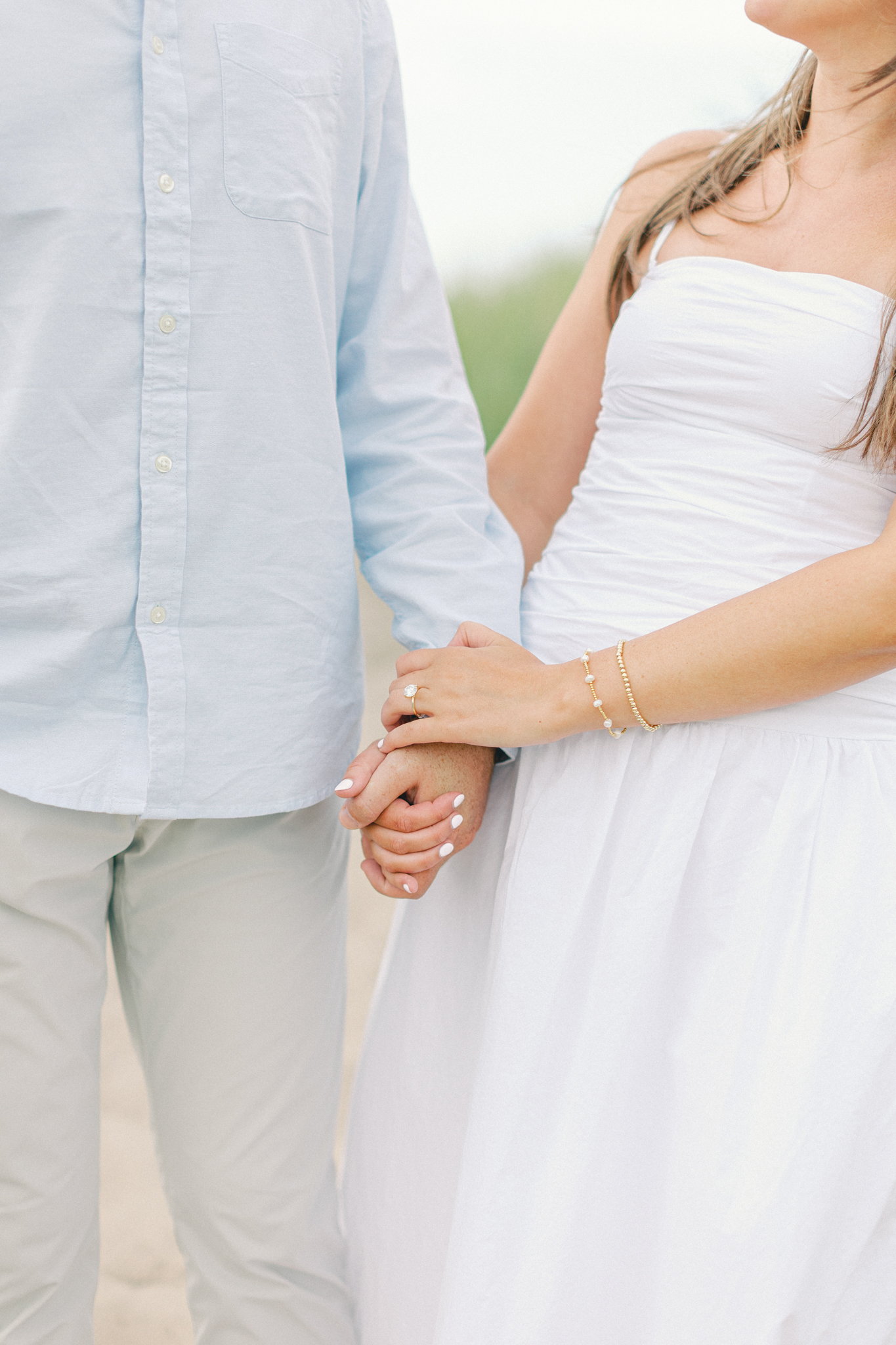 Close-up of an engaged couple holding hands, her in a white dress and him in a light blue shirt