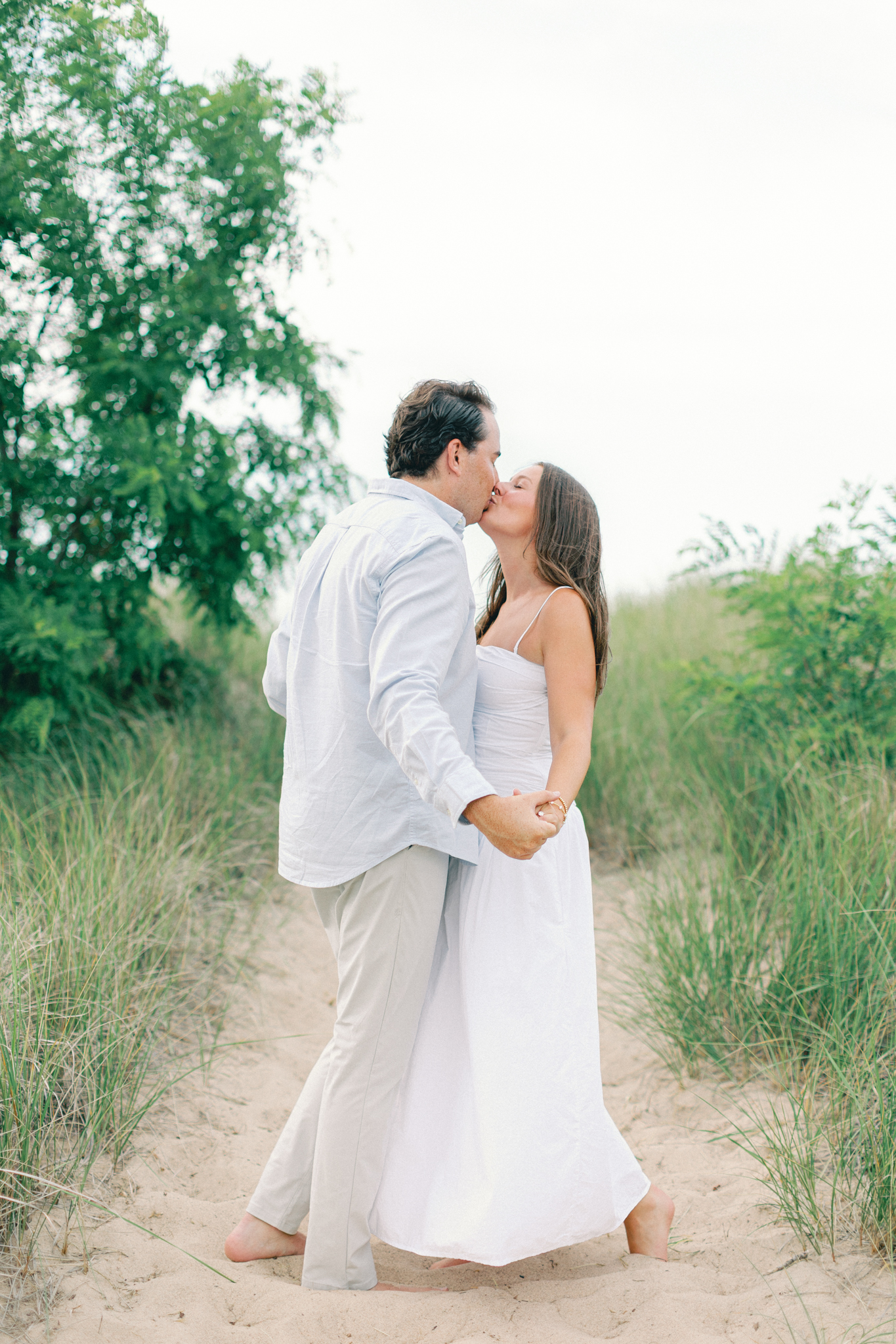 Lauren and Jason kissing on a sandy path lined with trees and dune grass