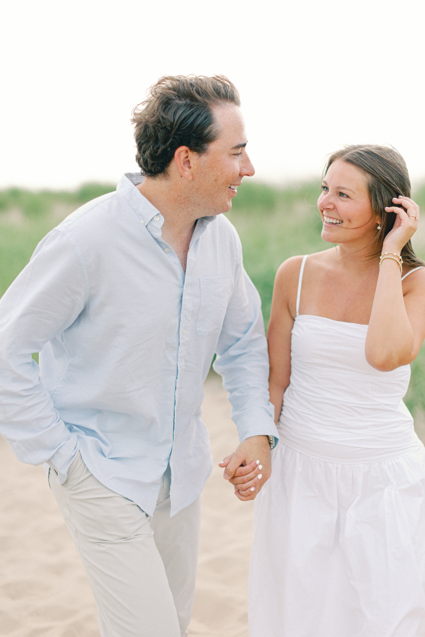 Lauren tucking her hair back and smiling at Jason as they walk hand in hand on a Lake Michigan beach