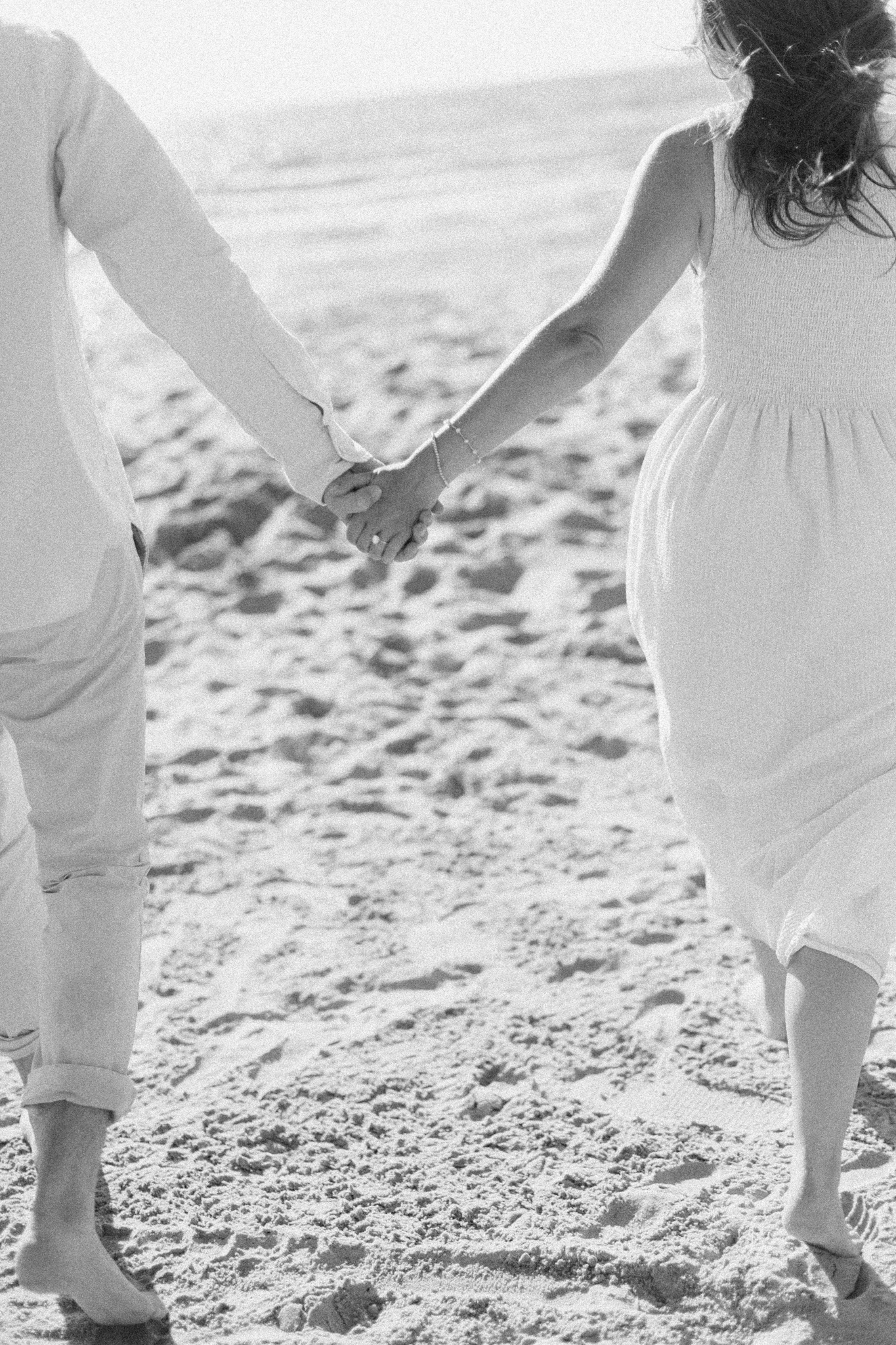 Black and white detail of an engaged couple holding hands as they walk across the sand