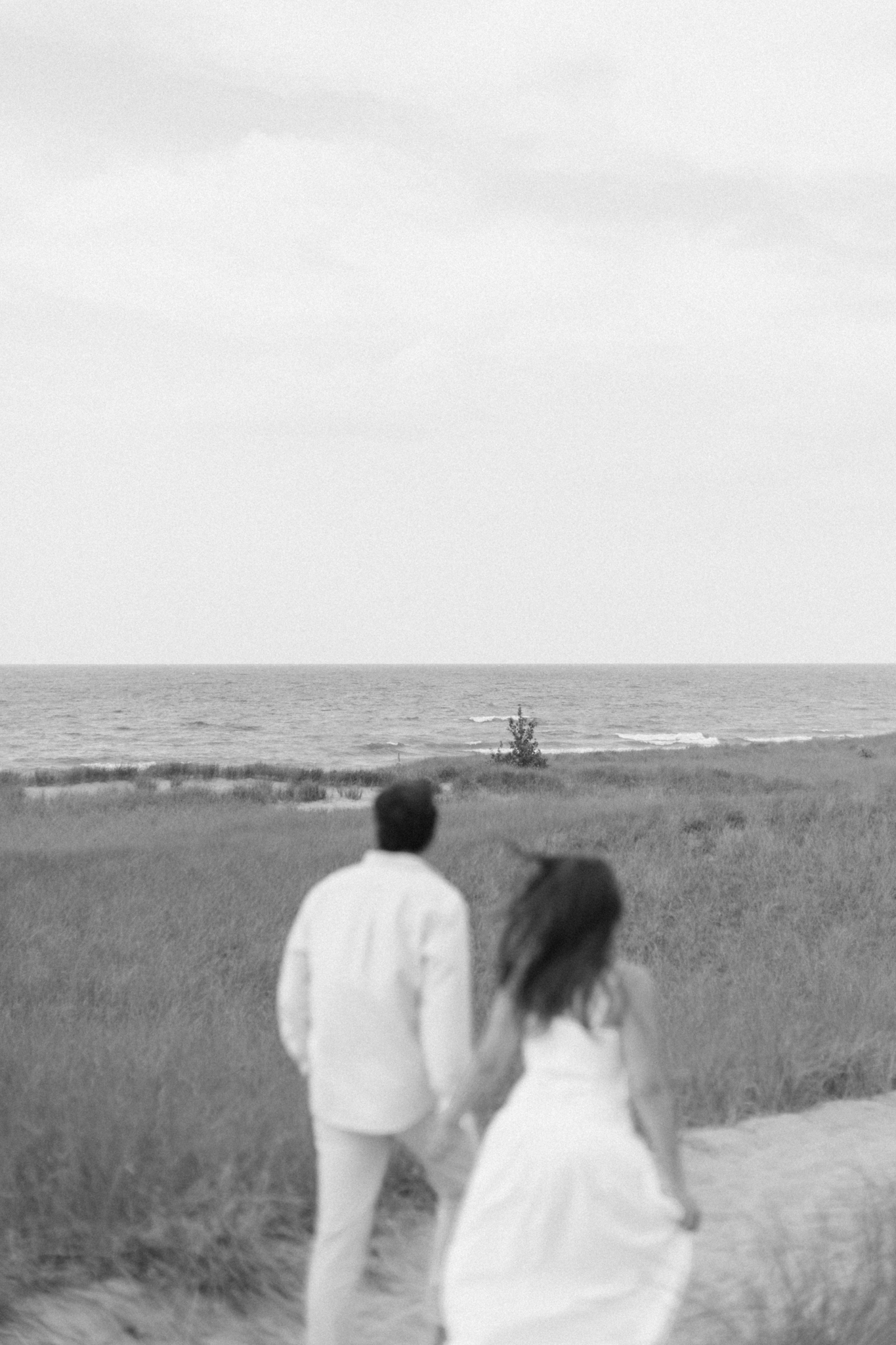Black and white photo of Lauren and Jason walking through dune grass toward Lake Michigan