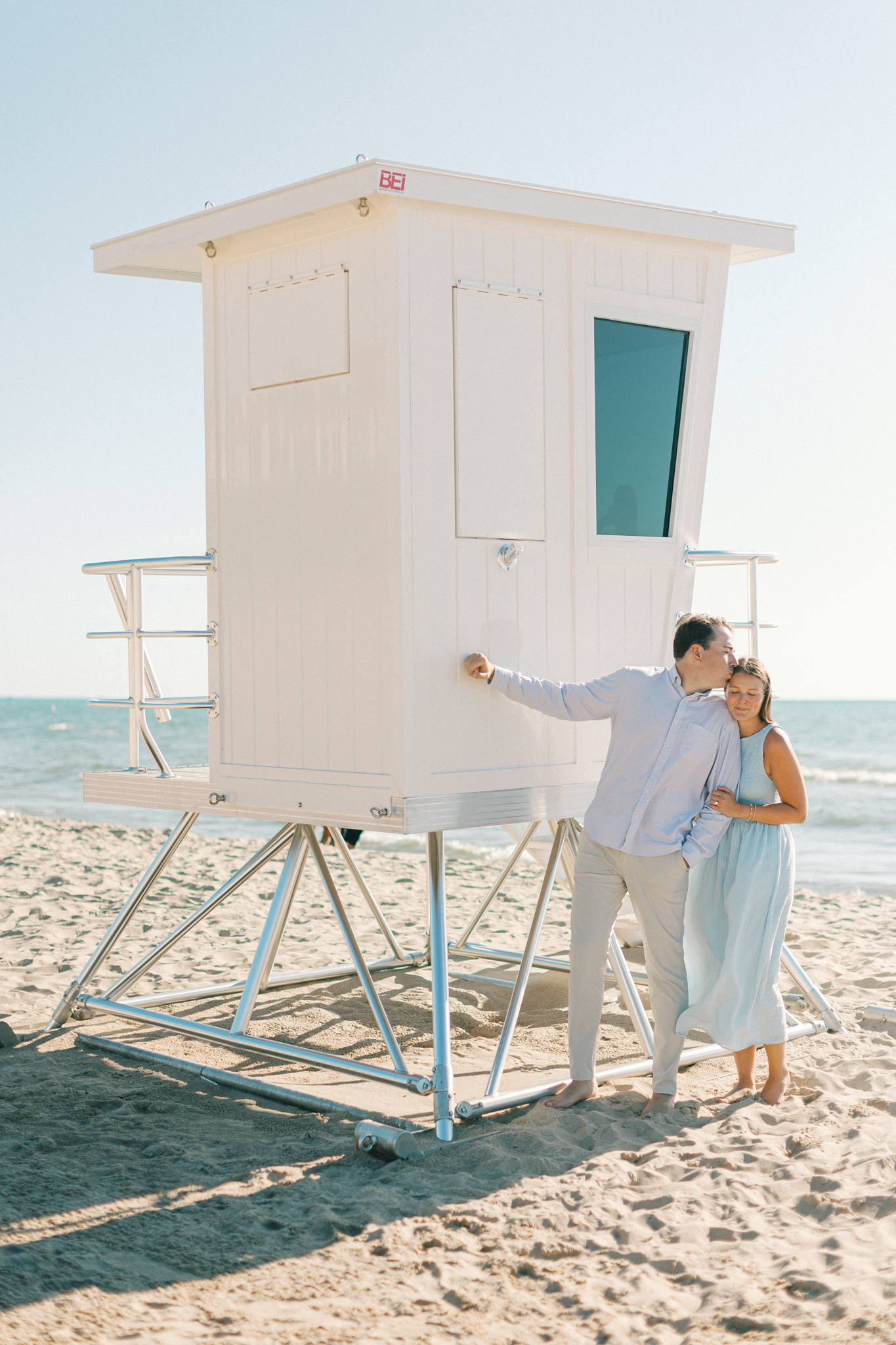 Lauren and Jason kissing beside a white lifeguard stand on a South Haven beach