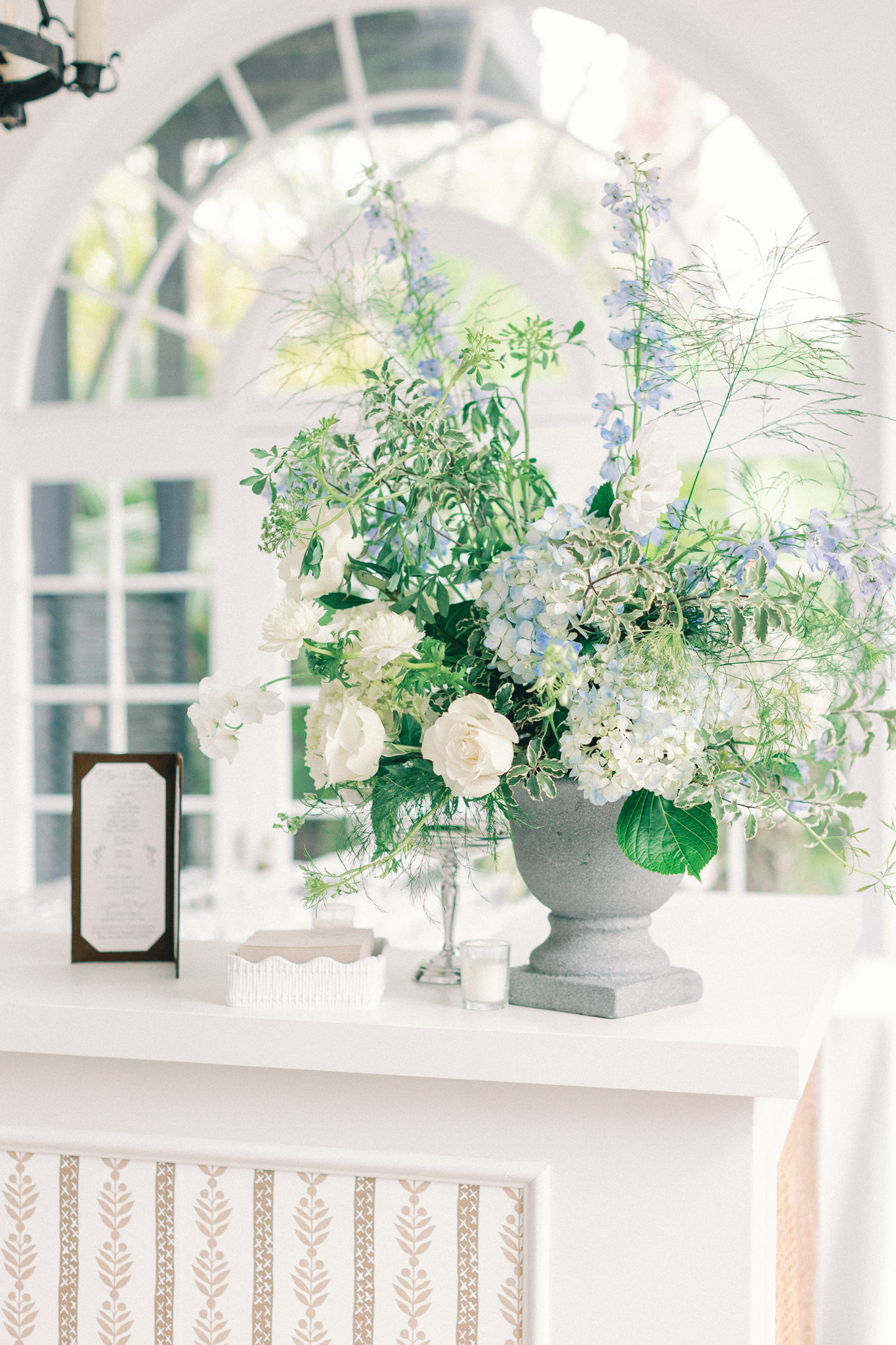 Blue and white hydrangea and delphinium in a stone urn on a mantel beneath an arched window