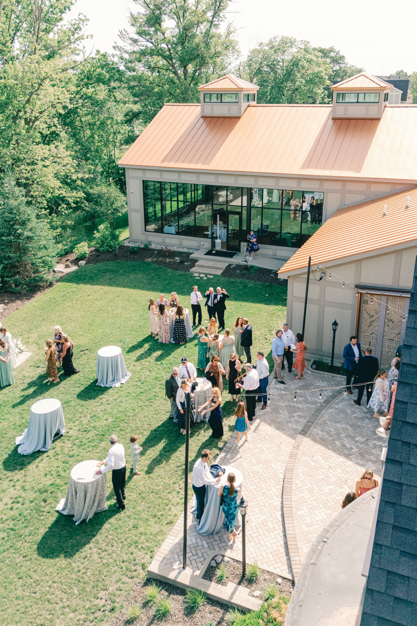 Aerial view of guests at cocktail hour on a lawn beside a glass-walled reception hall at a northern Michigan summer wedding