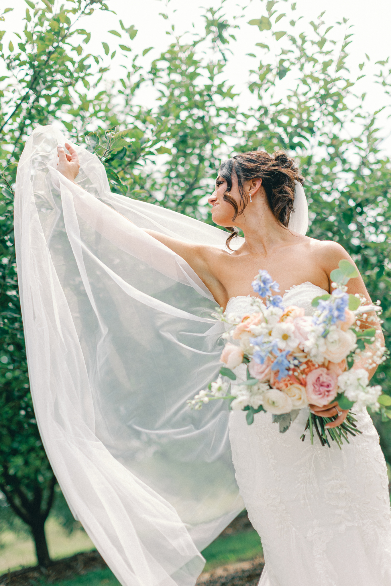Bride lifting her veil in an orchard while holding a peach and blue bouquet, Michigan wedding photos