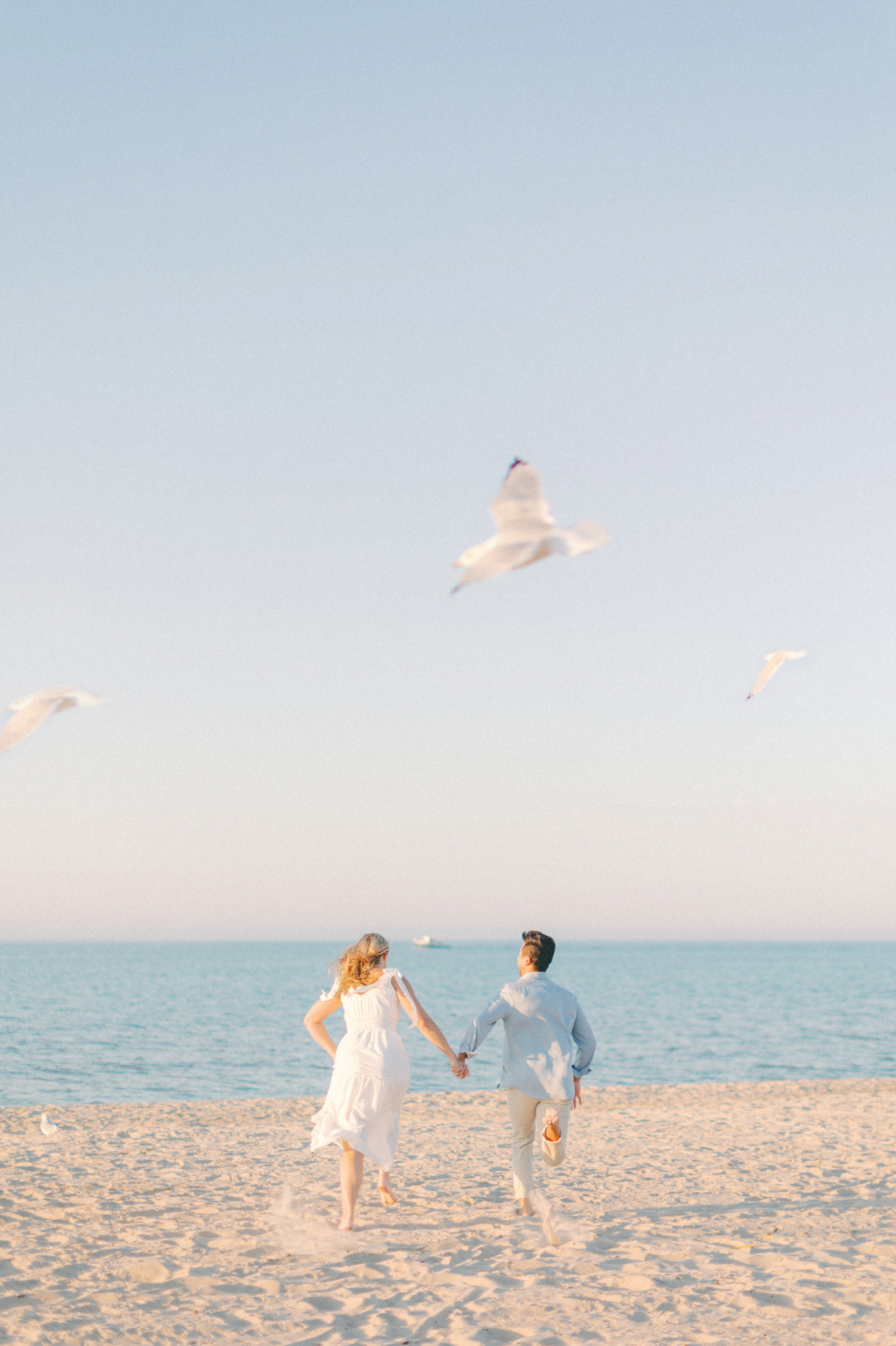 Haley and Andrew run toward the water as gulls fly overhead in Michigan City, photographed by a Michigan engagement photographer