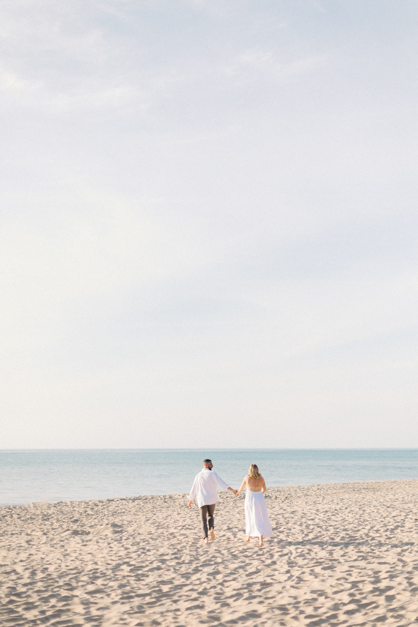 Couple walking hand in hand toward the water on a wide Lake Michigan beach, Michigan engagement photography