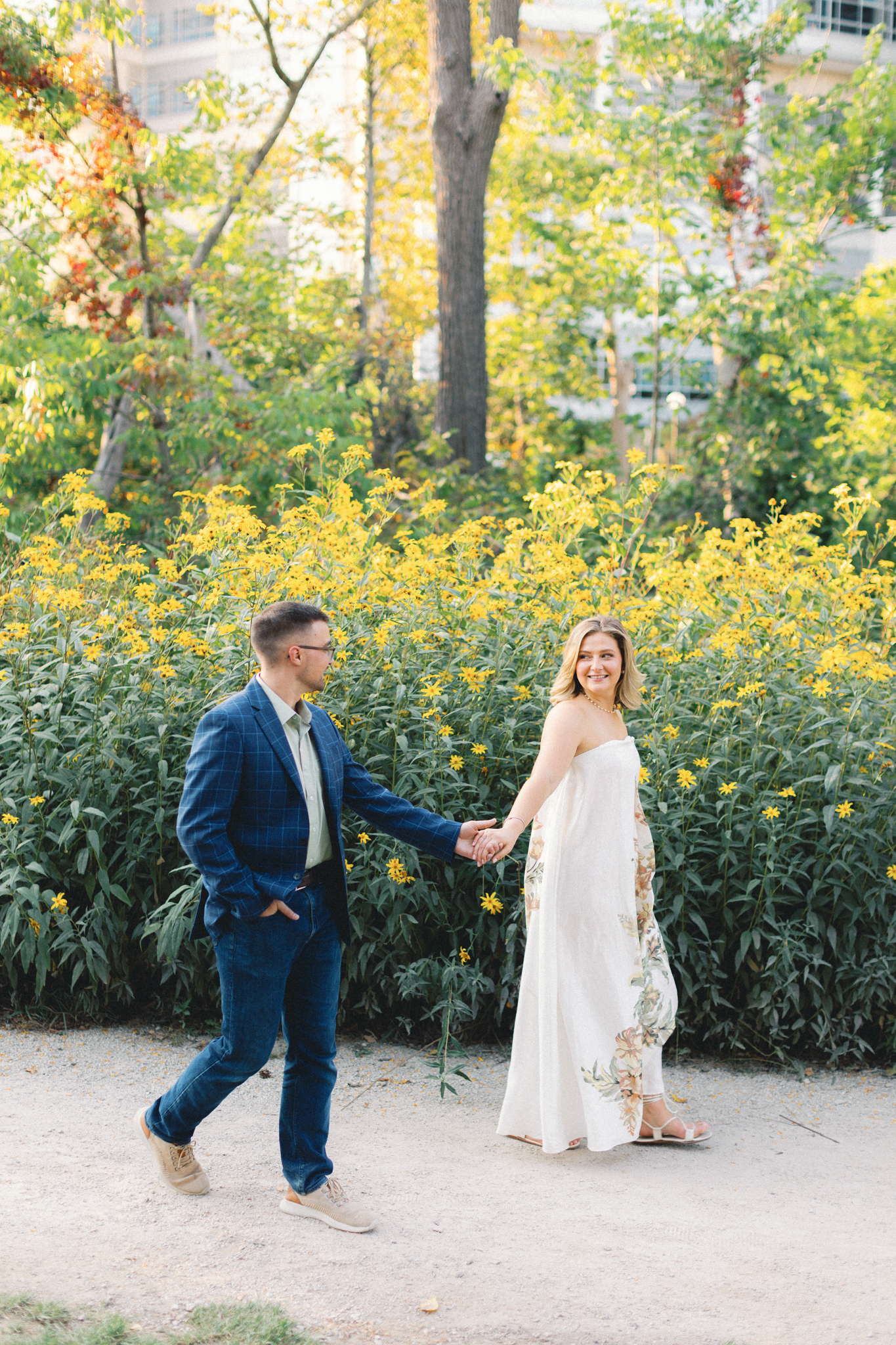 Melissa and David walk hand in hand past yellow wildflowers during their Ann Arbor engagement photos
