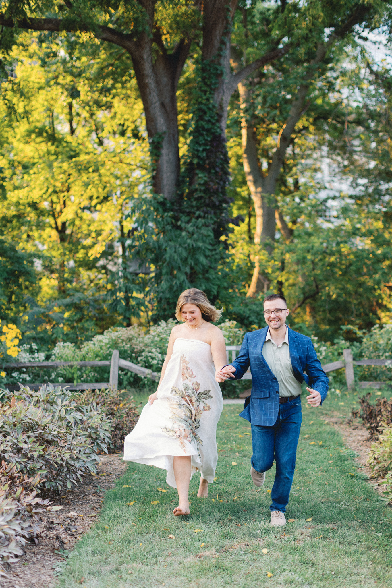 Melissa and David run hand in hand through the peony garden at Nichols Arboretum, photographed by a Michigan engagement photographer