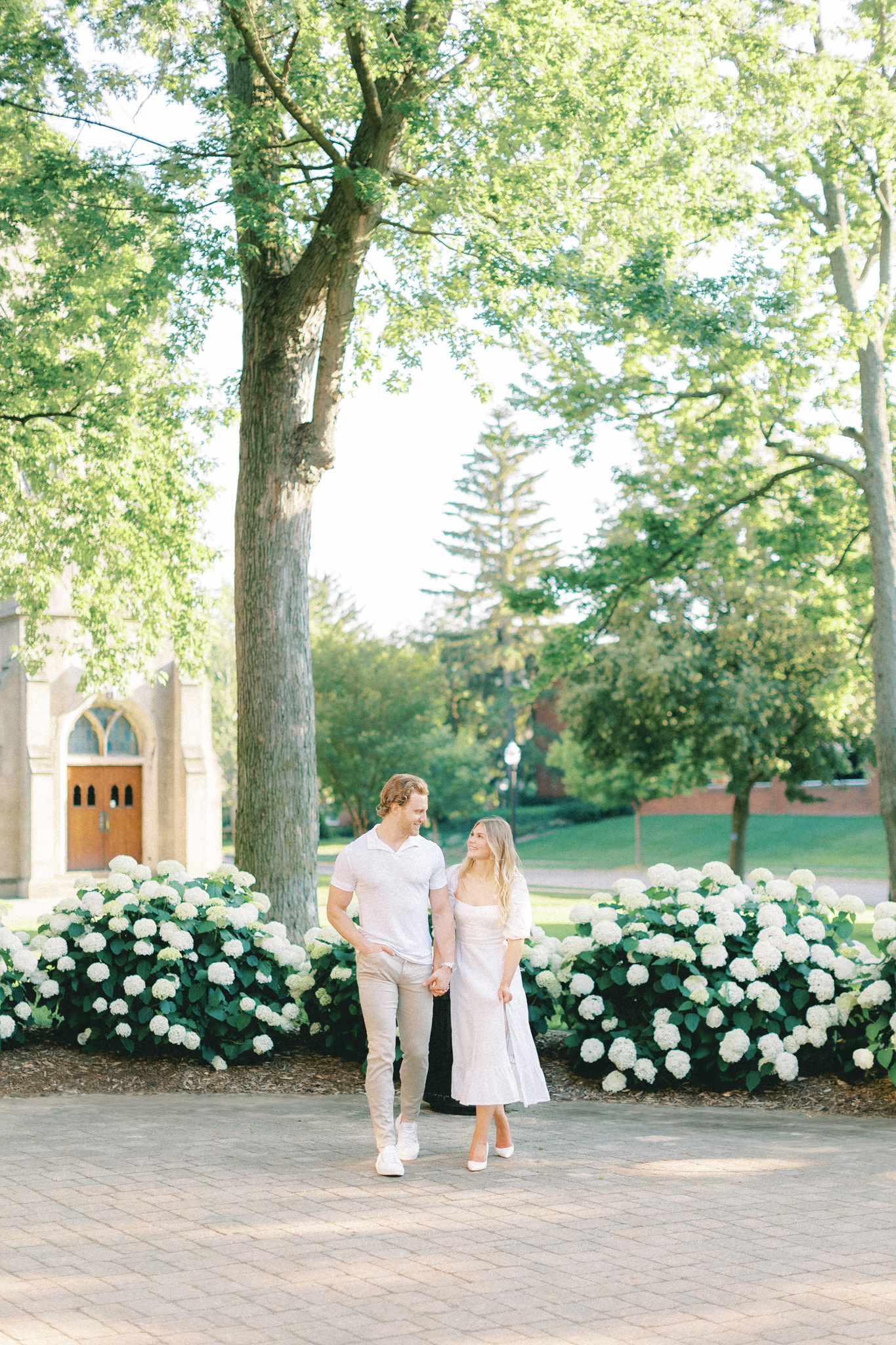 Claire and Patrick walk past white hydrangeas on the Hope College campus during their West Michigan engagement photos