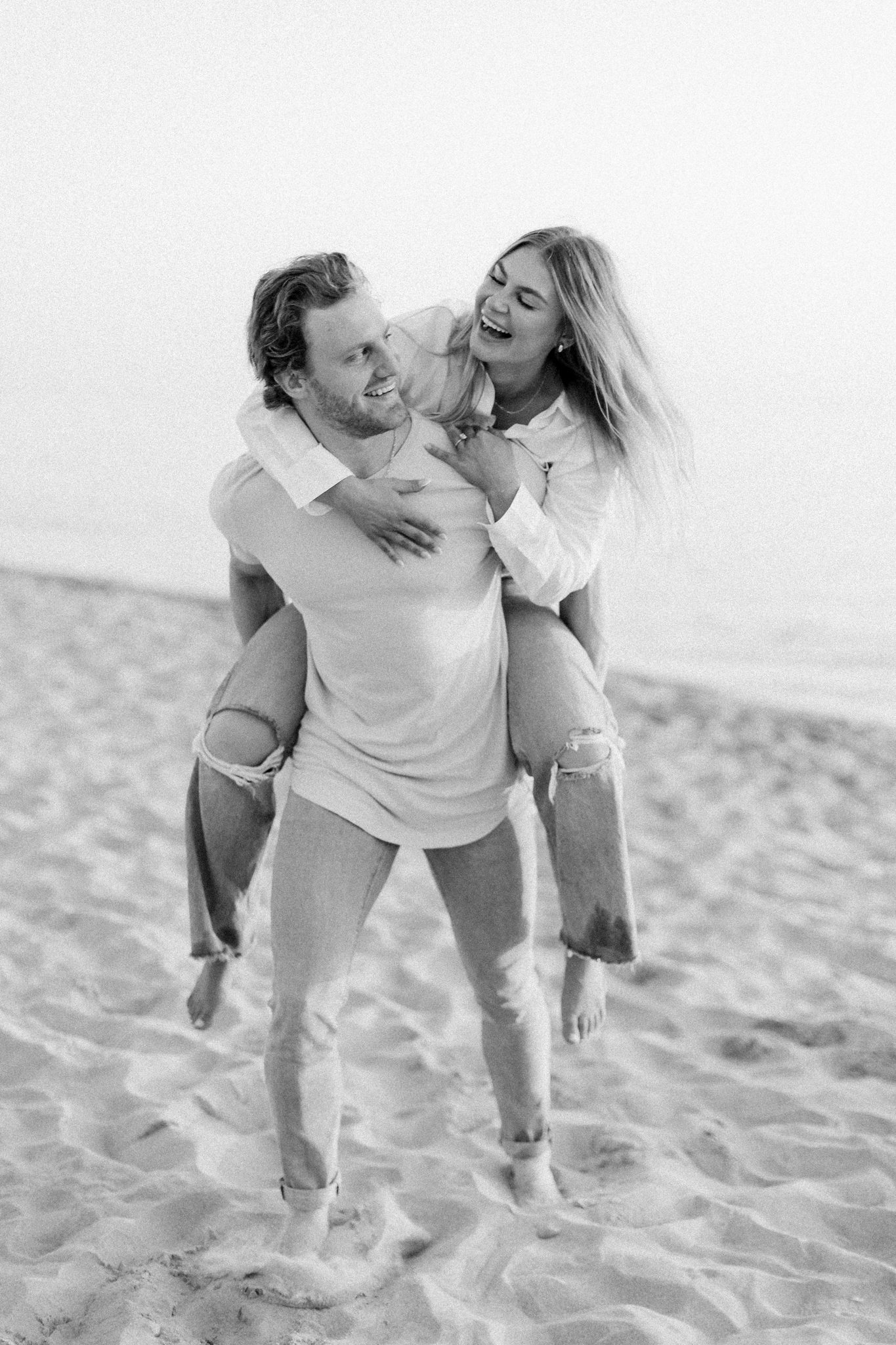 Black and white photo of Claire and Patrick laughing during a piggyback ride at Tunnel Park, West Michigan engagement photos in Holland