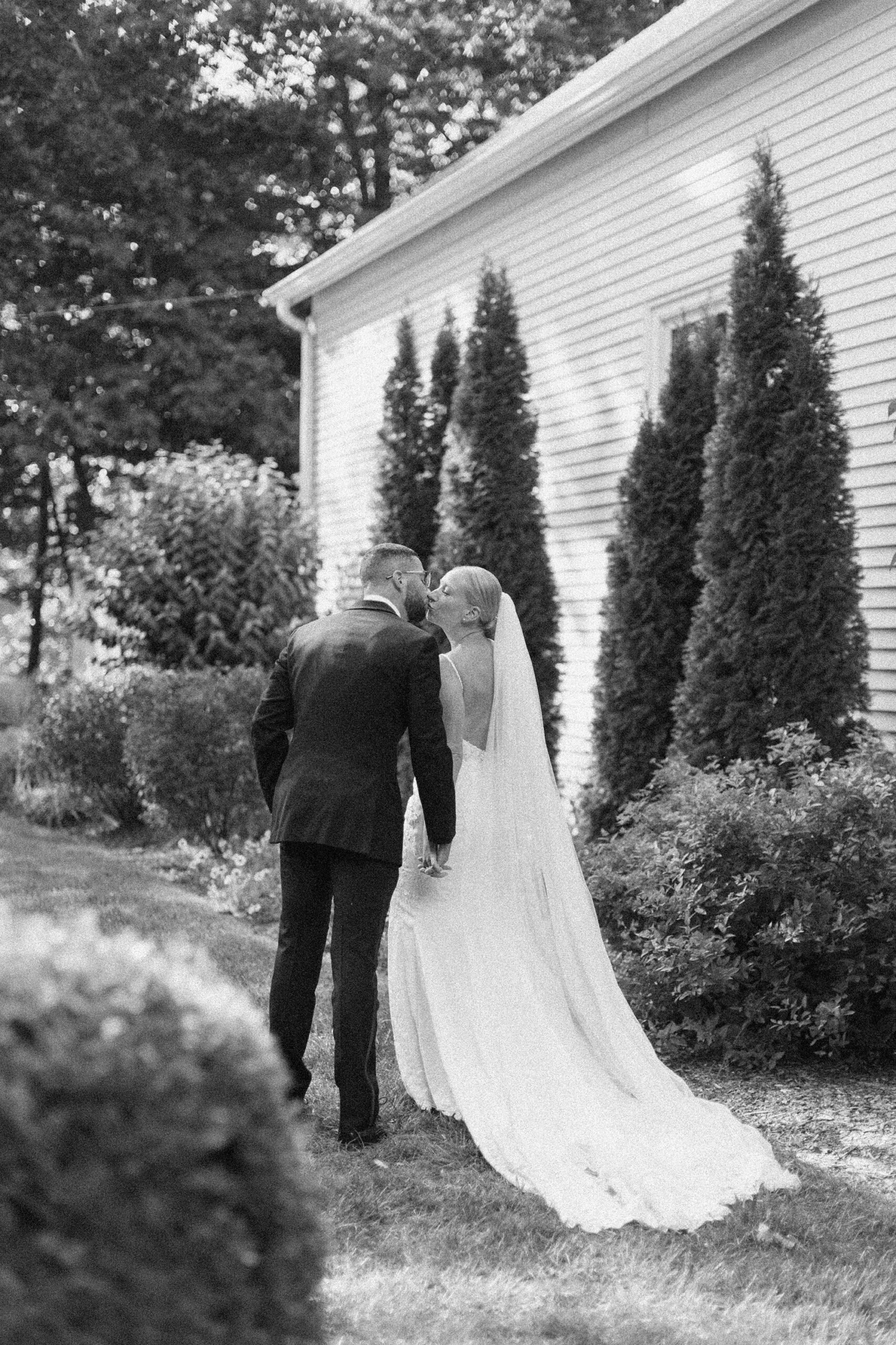 Bride and groom share a kiss beside a white cottage and tall hedges — northern Michigan wedding photography