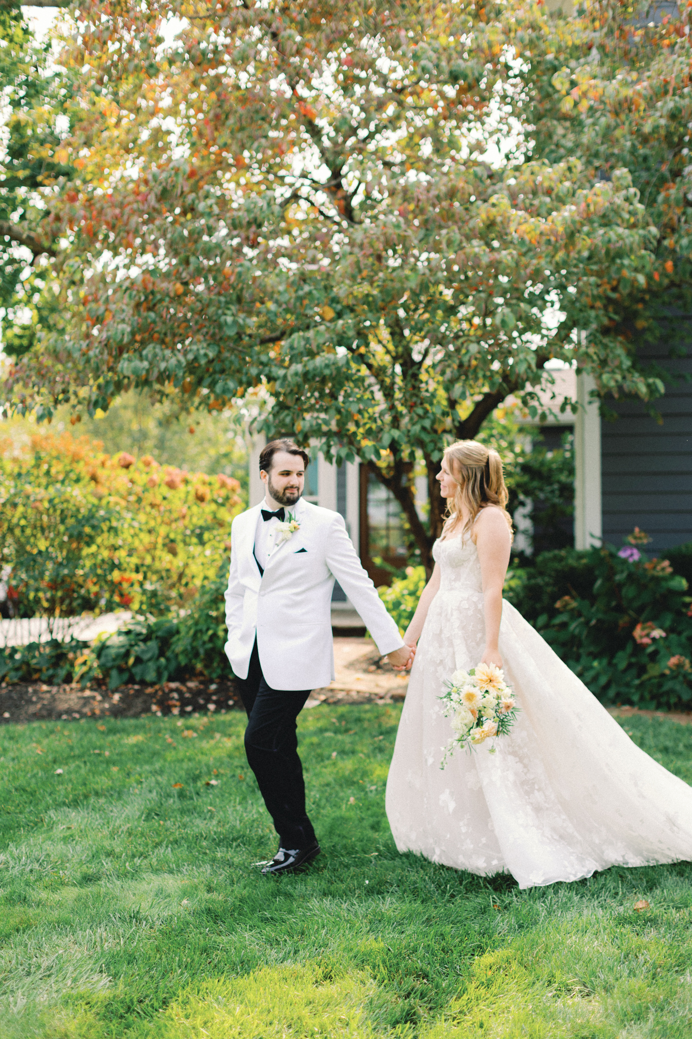 Bride and groom walking hand in hand under a tree turning red and orange, photographed by a Michigan wedding photographer